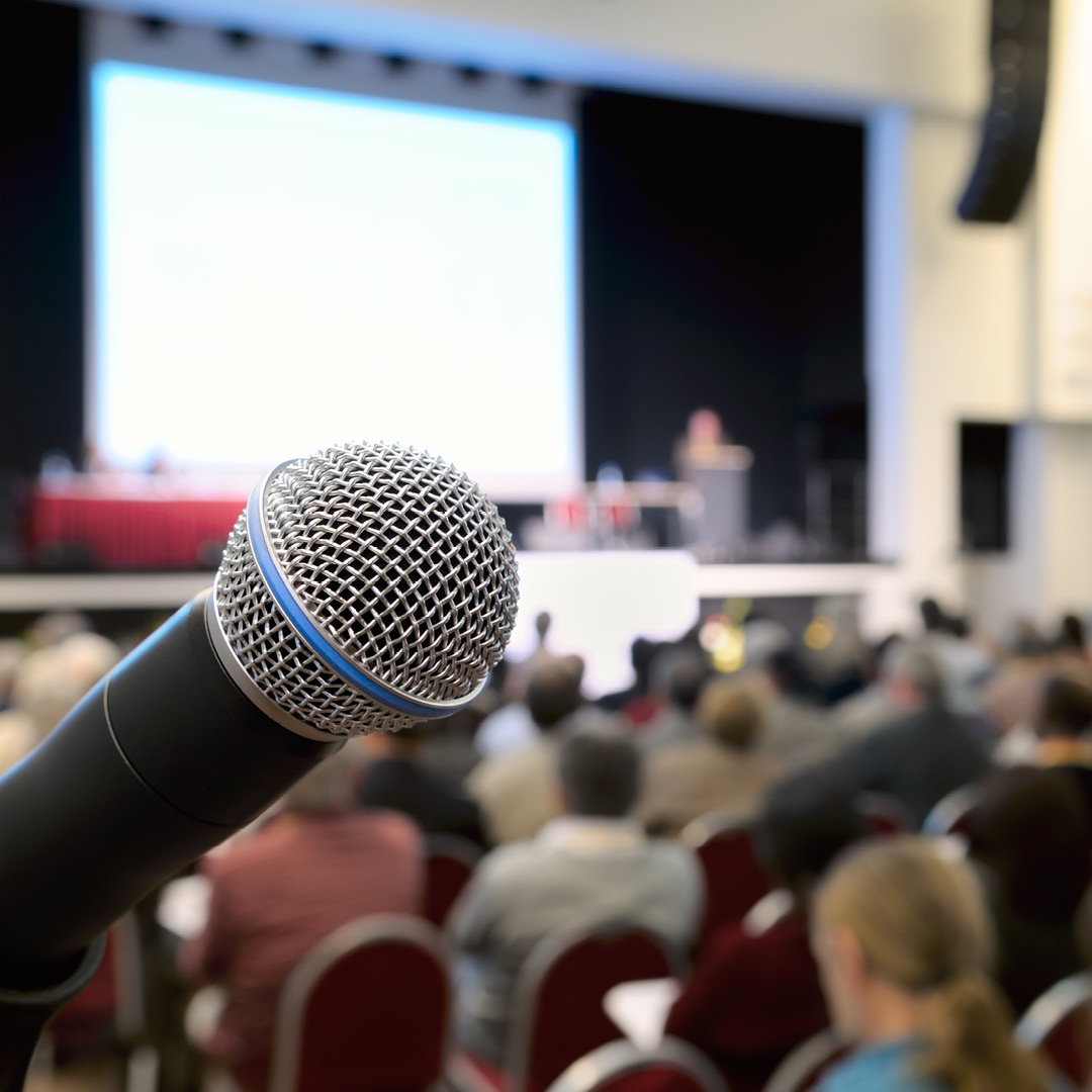 Un microfono sulla sinistra si staglia su un'affollata aula magna, sul palco si tiene una conferenza