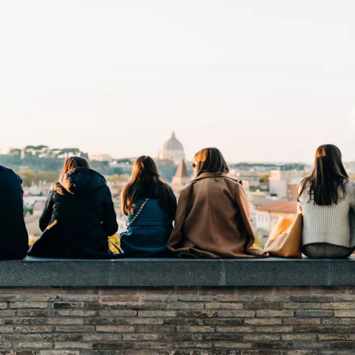 Nella foto si vedono cinque persone sedute di spalle su un muretto di pietra. Davanti a loro si apre un panorama urbano illuminato dalla luce del tramonto. Sullo sfondo si riconosce una grande cupola, circondata da edifici e alberi. Le persone indossano giacche e cappotti, come in una giornata autunnale o invernale, e sembrano godersi la vista in un momento di quiete.