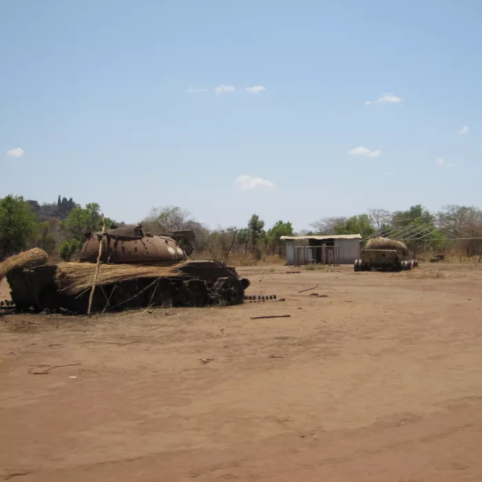Nell’immagine si vede un terreno arido e polveroso sotto un cielo azzurro chiaro. In primo piano ci sono due vecchi veicoli corazzati arrugginiti, parzialmente coperti da erba secca o paglia. Sullo sfondo si intravedono alcuni alberi sparsi e una piccola costruzione semplice, probabilmente in lamiera. L’ambiente appare rurale e tranquillo, con un’atmosfera di abbandono.