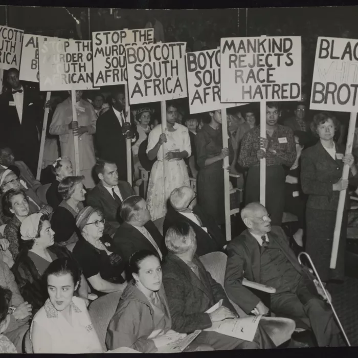 L’immagine ritrae un’assemblea o manifestazione al chiuso, con molte persone sedute e altre in piedi.
